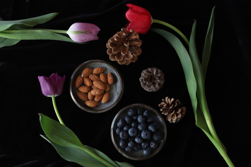 Overhead still life with three tulips and three pine cones arranged around two dishes, one with about a handful of almonds and one with about 30 blueberries all on a dark background.