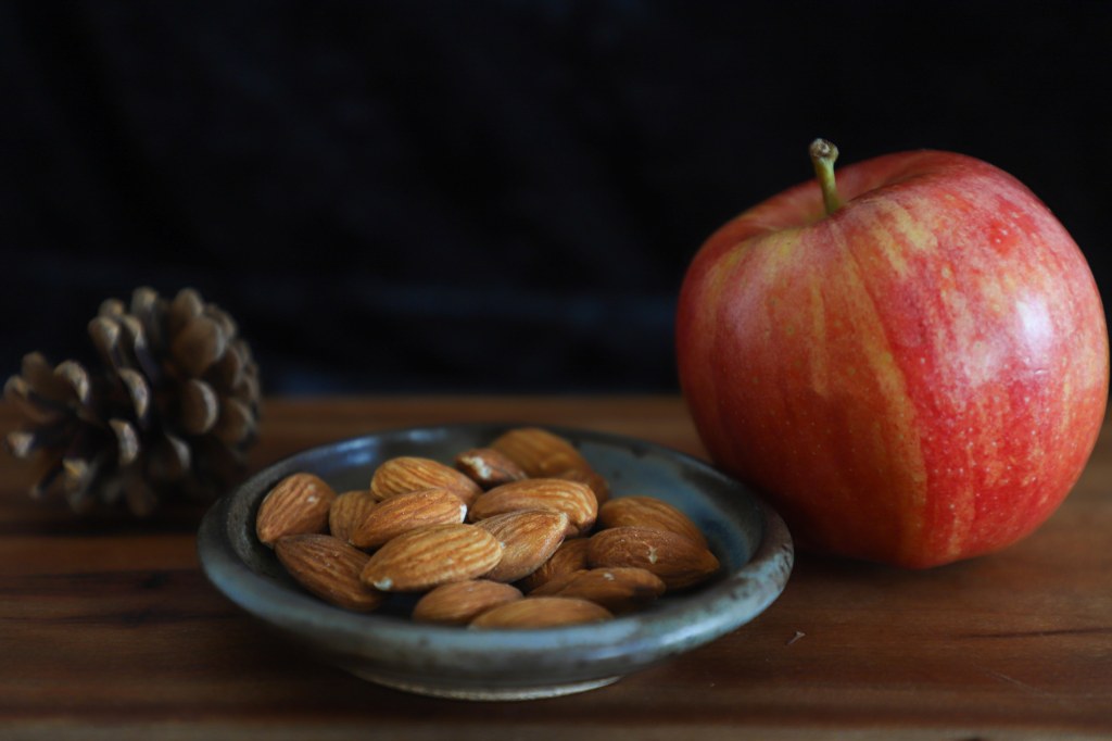 shallow dish of almonds on a wooden surface, pinecone in the background on the left, apple beside on the right.