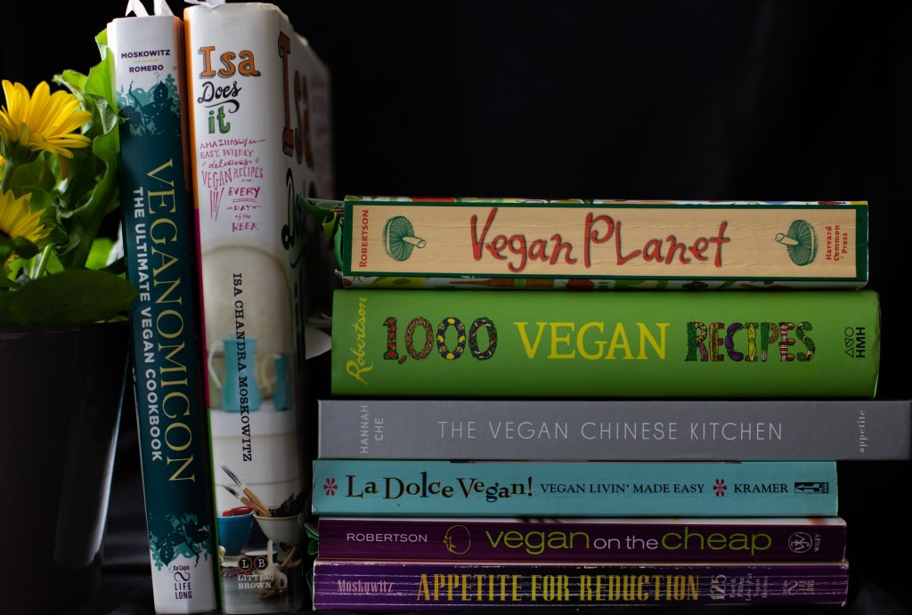 Arrangement of eight vegan cookbooks and a gerber daisy plant to their left. Upright cookbooks on the left: Veganomicon and Isa Does It. Cookbooks stacked horizontally on the right side of those two, from top to bottom: Vegan Planet, 1000 Vegan Recipes, The Vegan Chinese Kitchen, La Dolce Vegan!, vegan on the cheap, Appetite for Reduction.