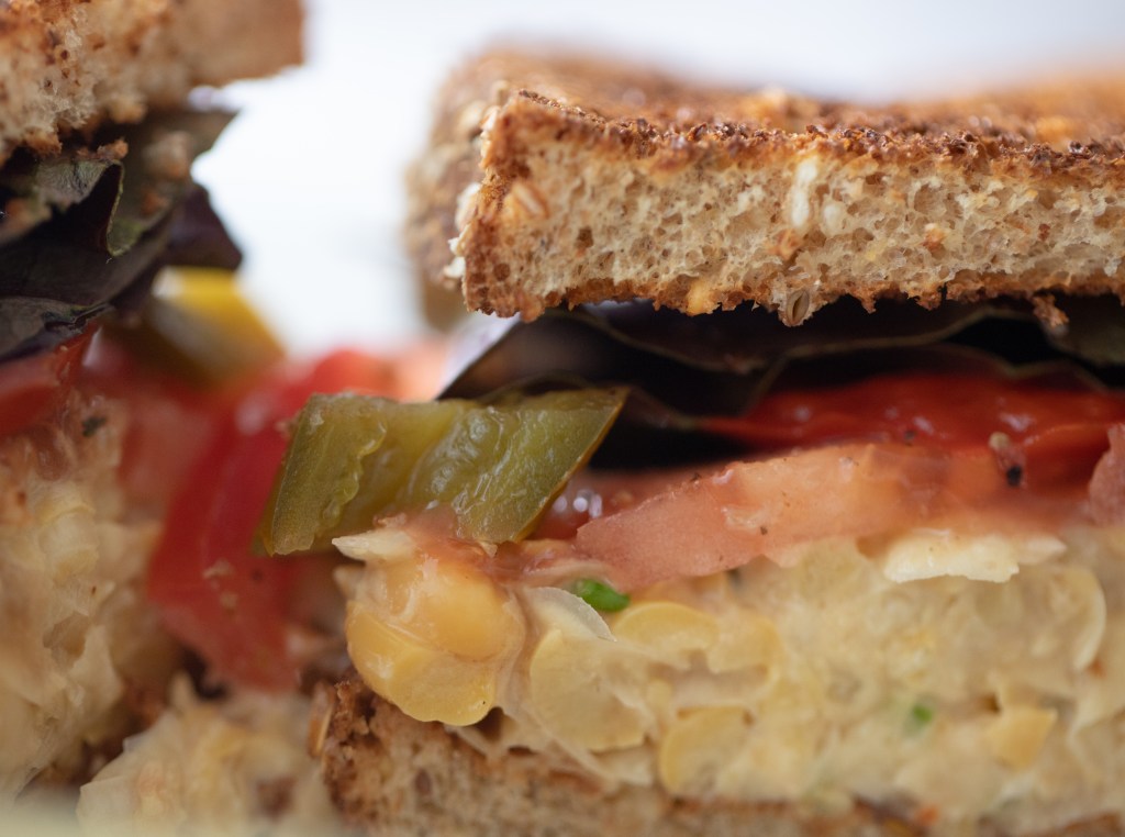 Close up of a chickpea salad sandwich on whole wheat toast with sliced tomato, pickled jalapeno, and lettuce.