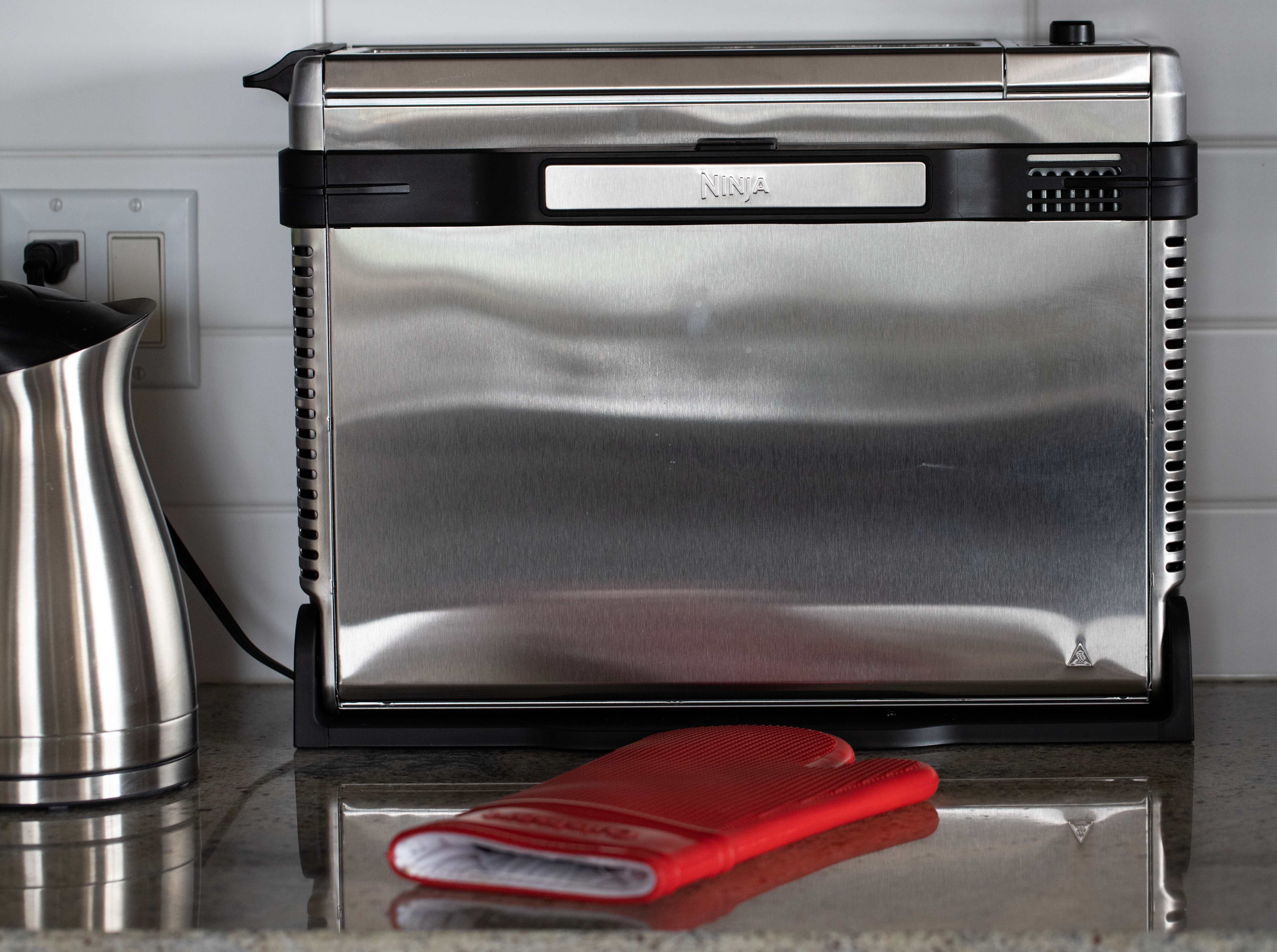 Stainless steel air fryer, in the flipped up position, with a stainless electric kettle beside it and a red silicone oven mitt in front on a granite countertop with a white tile backsplash.