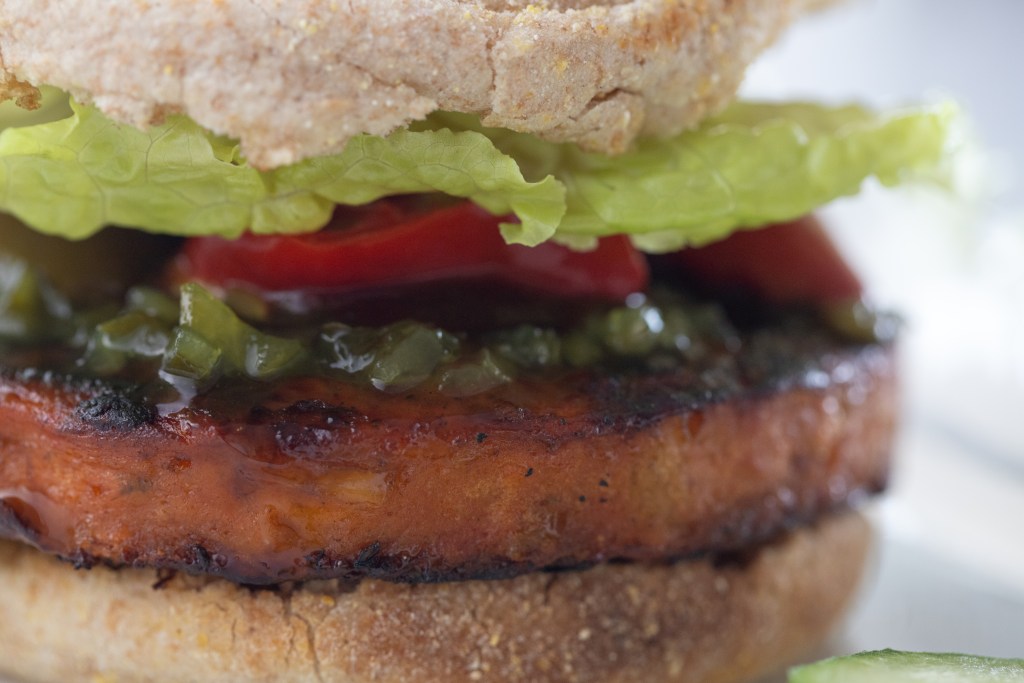 Close up of a veggie burger with relish, red banana peppers, and lettuce on a whole wheat English muffin, small slice of cucumber on the bottom right. (photo by Tracy Isaacs)