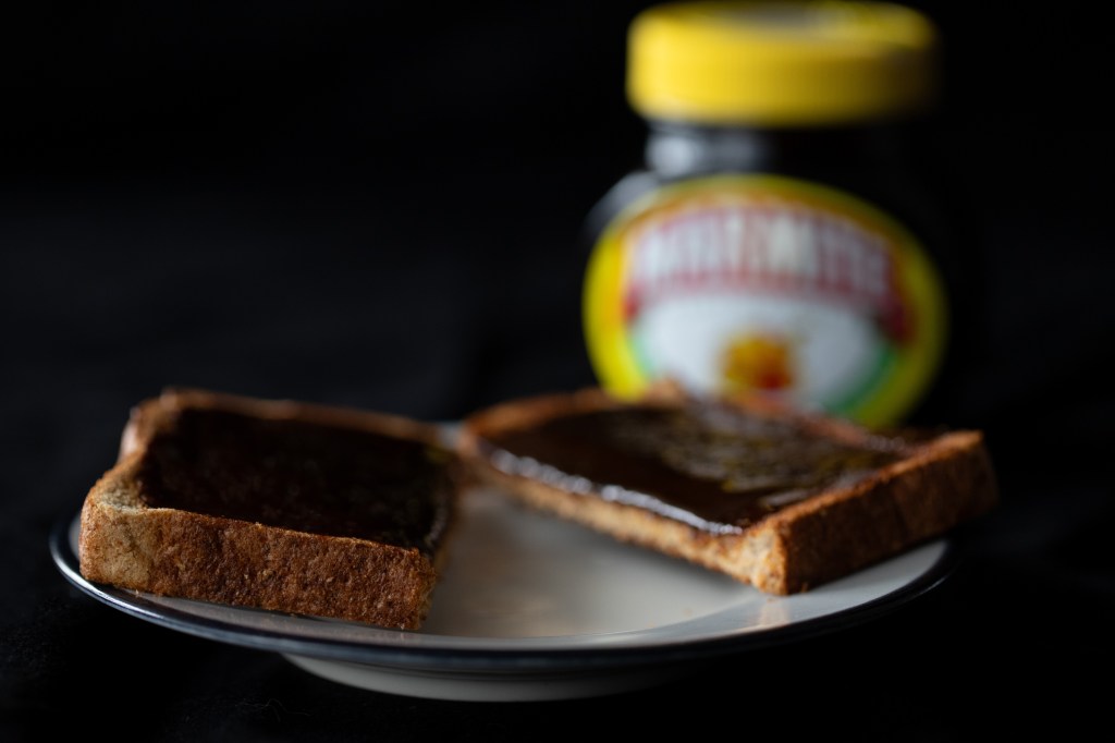 Close up of toast spread with Marmite (a black yeast spread), cut in half on a small plate, dark background, blurred jar of Marmite set in behind the plate. Photo by Tracy Isaacs