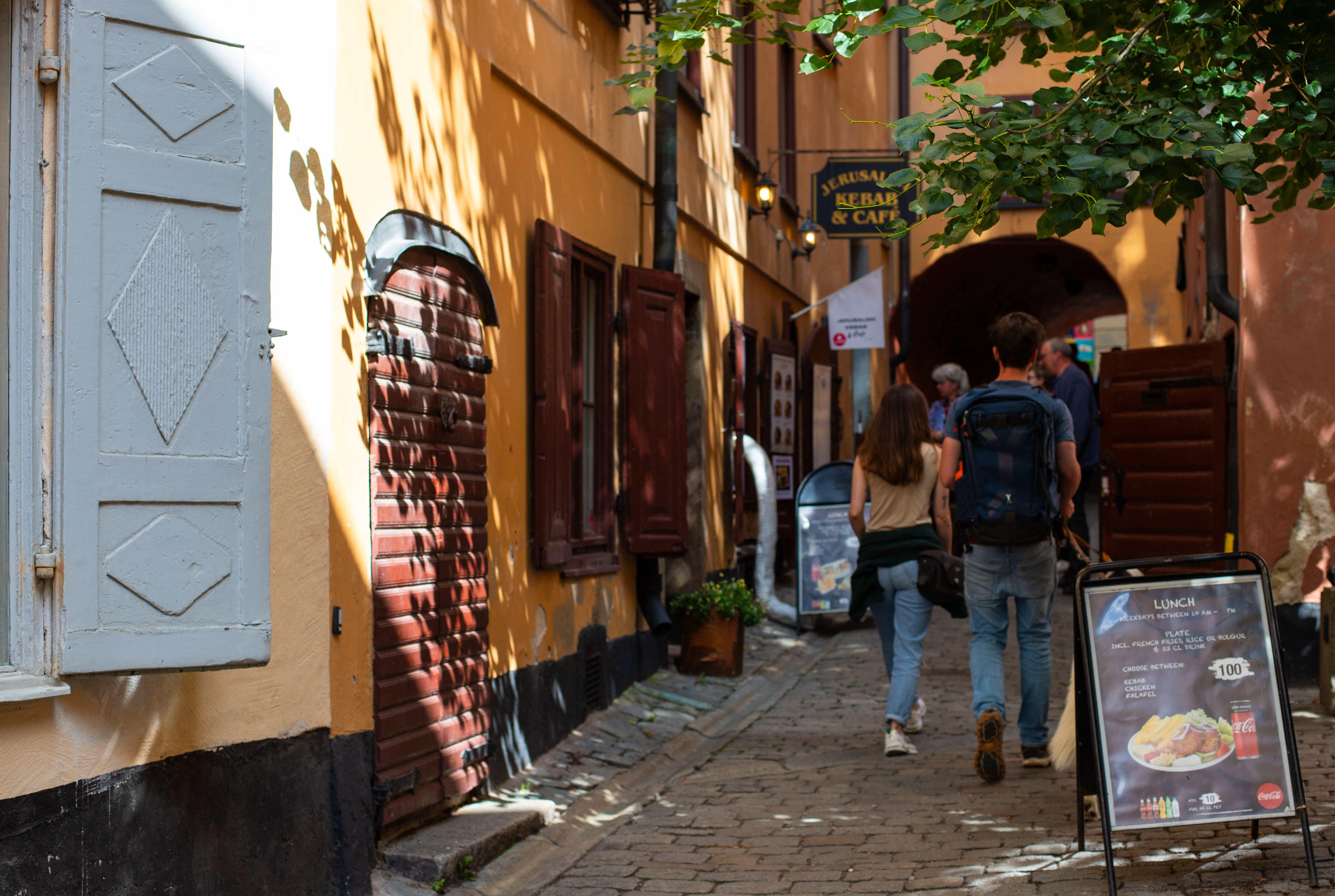 Outside narrow street scene in Old Stockholm (Gamla Stan) showing a brighly coloured alley and the front entrance of Jerusalem Kebab & Cafe on the left, stairs on the right, an advertising board on the cobblestoned street, a few people walking. Photo by Tracy Isaacs