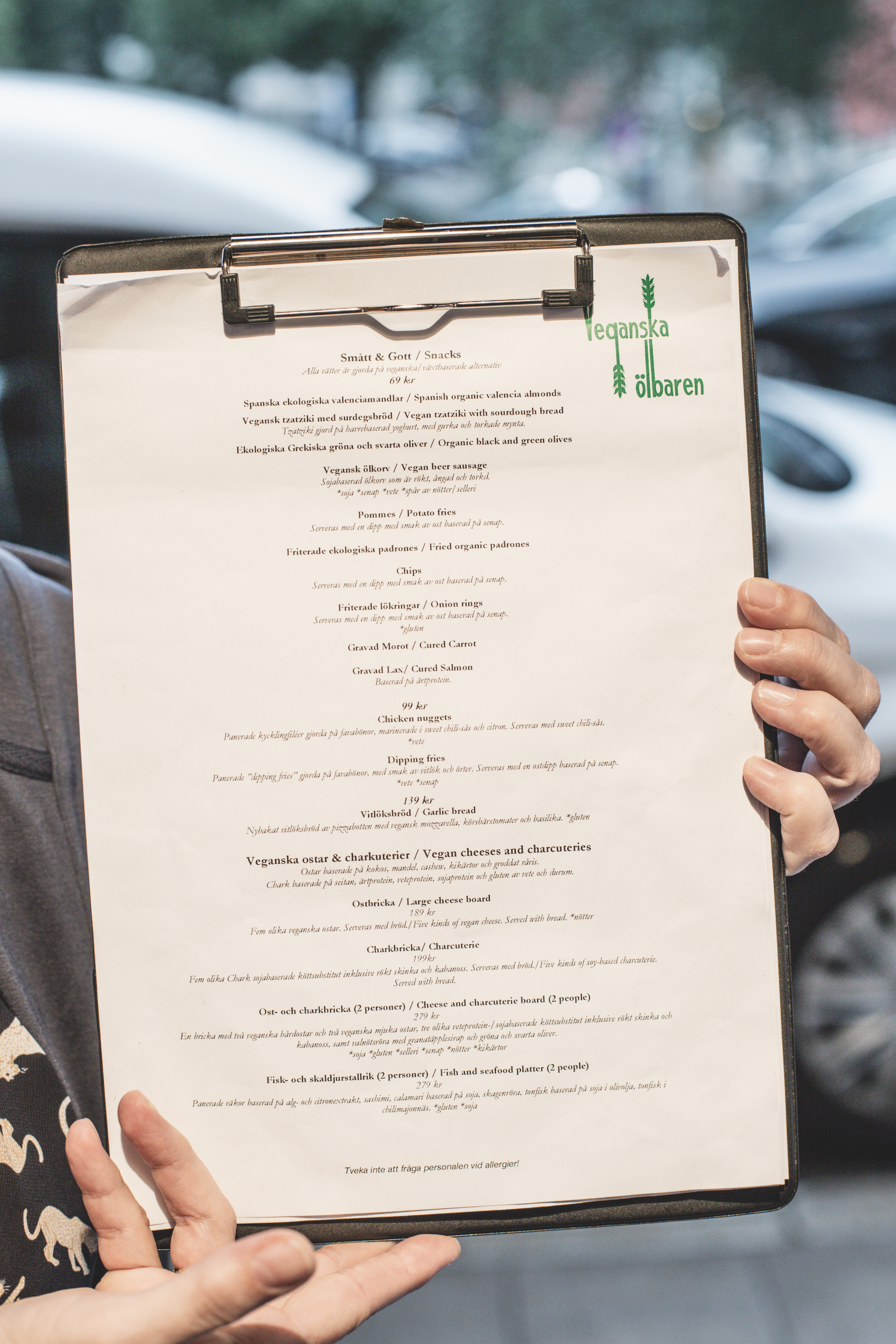 Close up someone holding a clipboard with the first page of the Veganska Olbaren menu on it. Blurred background of cars and buildings. Photo by Tracy Isaacs