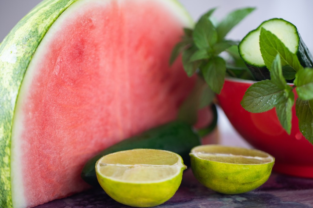 Ingredients for a watermelon salad including a cut watermelon, a sad looking lime cut in half, a jalapeno in behind the lime, a red bowl containing some freshly washed fresh mint and a piece of cucumber with the sliced side showing. Photo by Tracy Isaacs