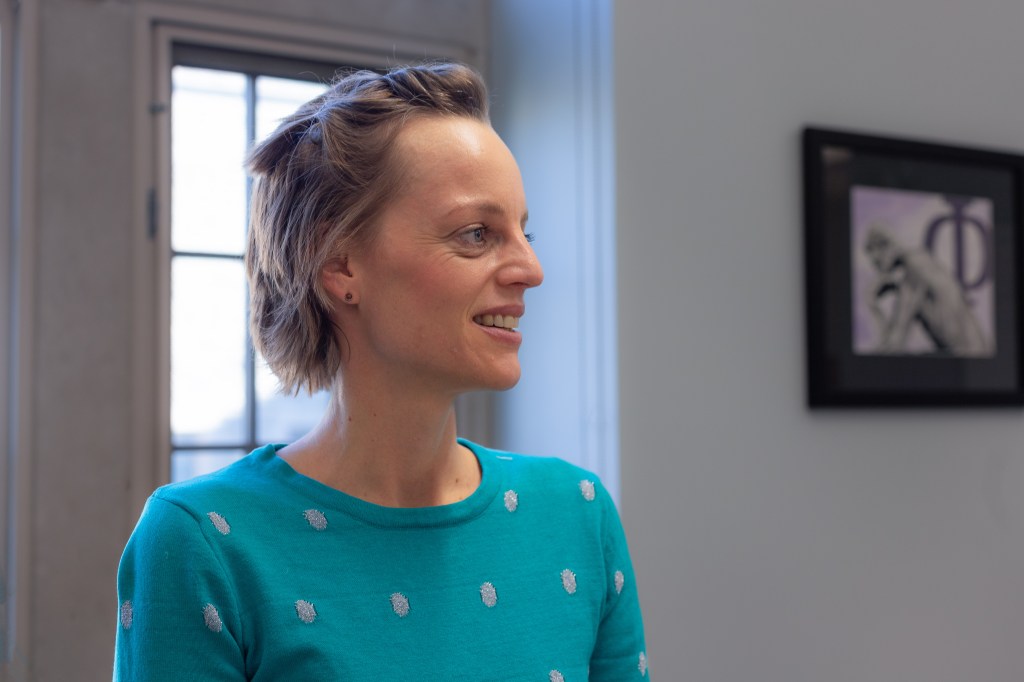 Profile of a young woman with chin-length hair pulled back, window and wall with artwork in the background. Photo by Tracy Isaacs