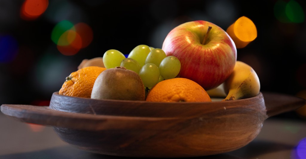 Shallow wooden fruit bowl containing kiwi, oranges, grapes, apple, and banana on a dark background and some blurred Christmas lights. Photo by Tracy Isaacs