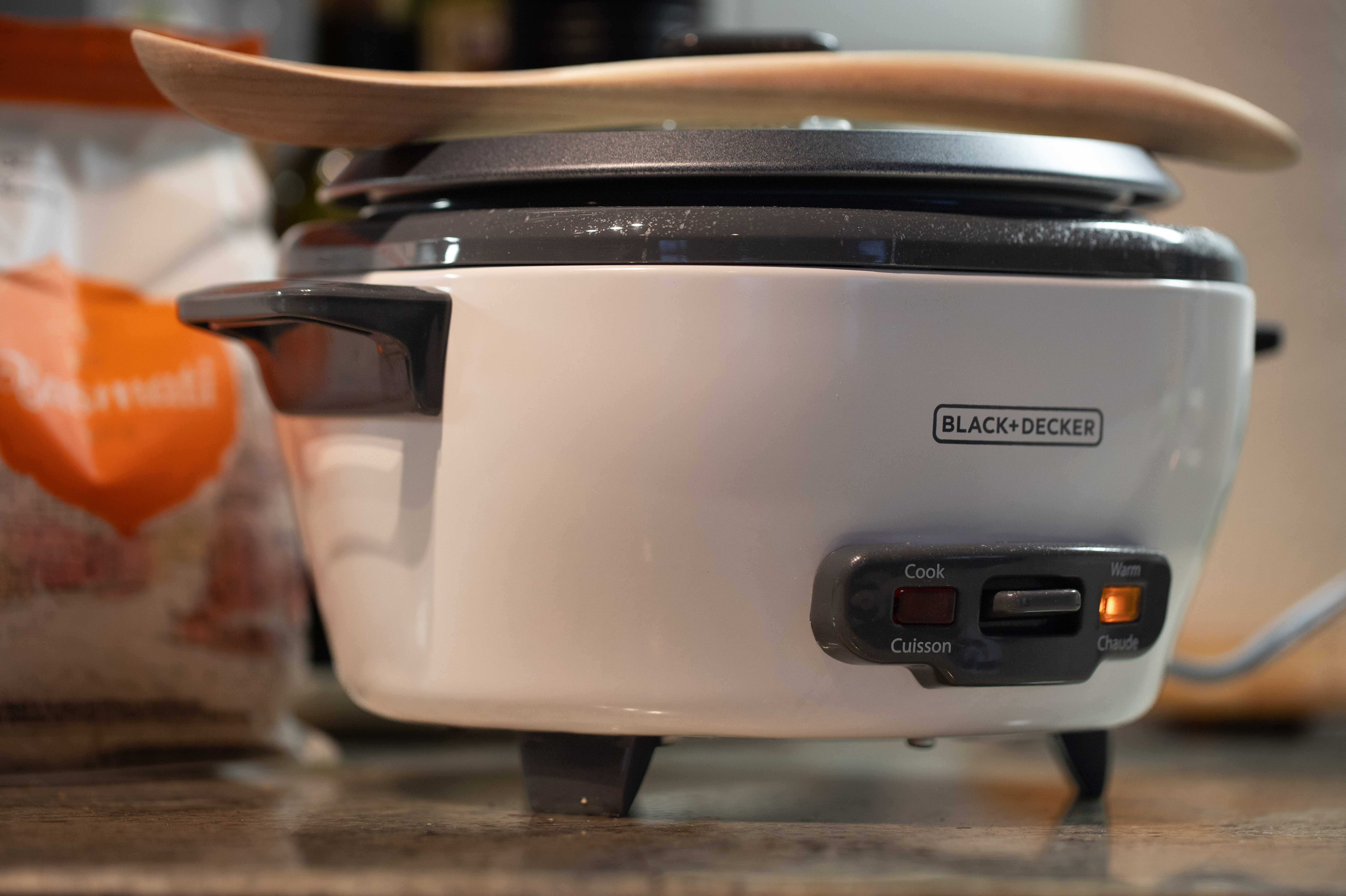 Black and Decker rice cooker, light on the warming setting, a wooden spoon resting atop the lid, bag of basmati in the background. Photo by Tracy Isaacs