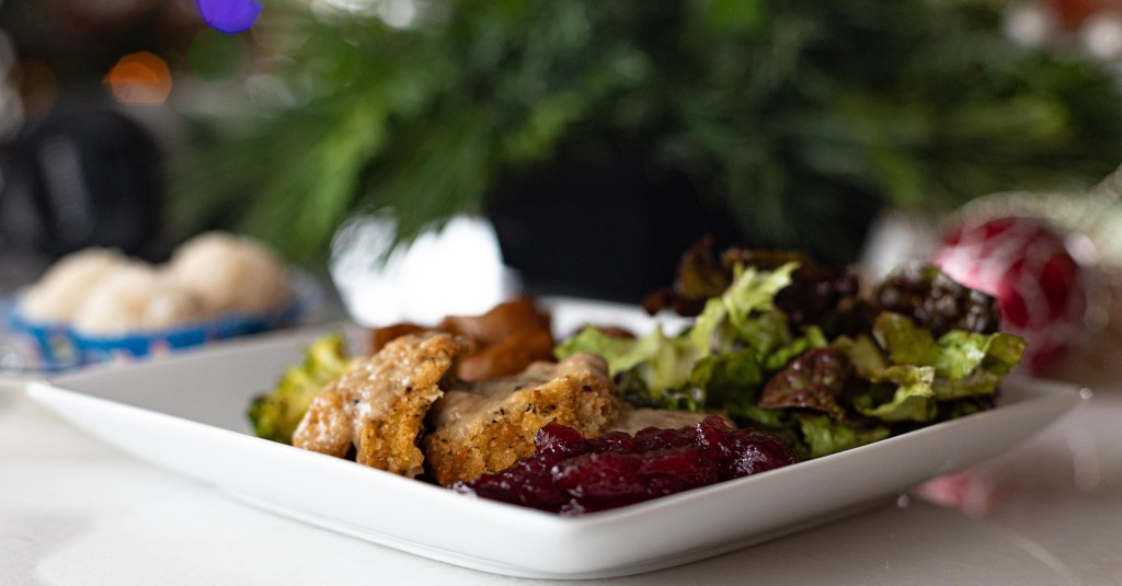 A square plate with a meal of Gardein stuffed "turk'y" sliced with gravy, cranberry sauce, salad and broccoli in the foreground and a blurred background with dessert on the left, a pine decorative plant in the centre, and a tree ornament on the right. Photo by Tracy Isaacs