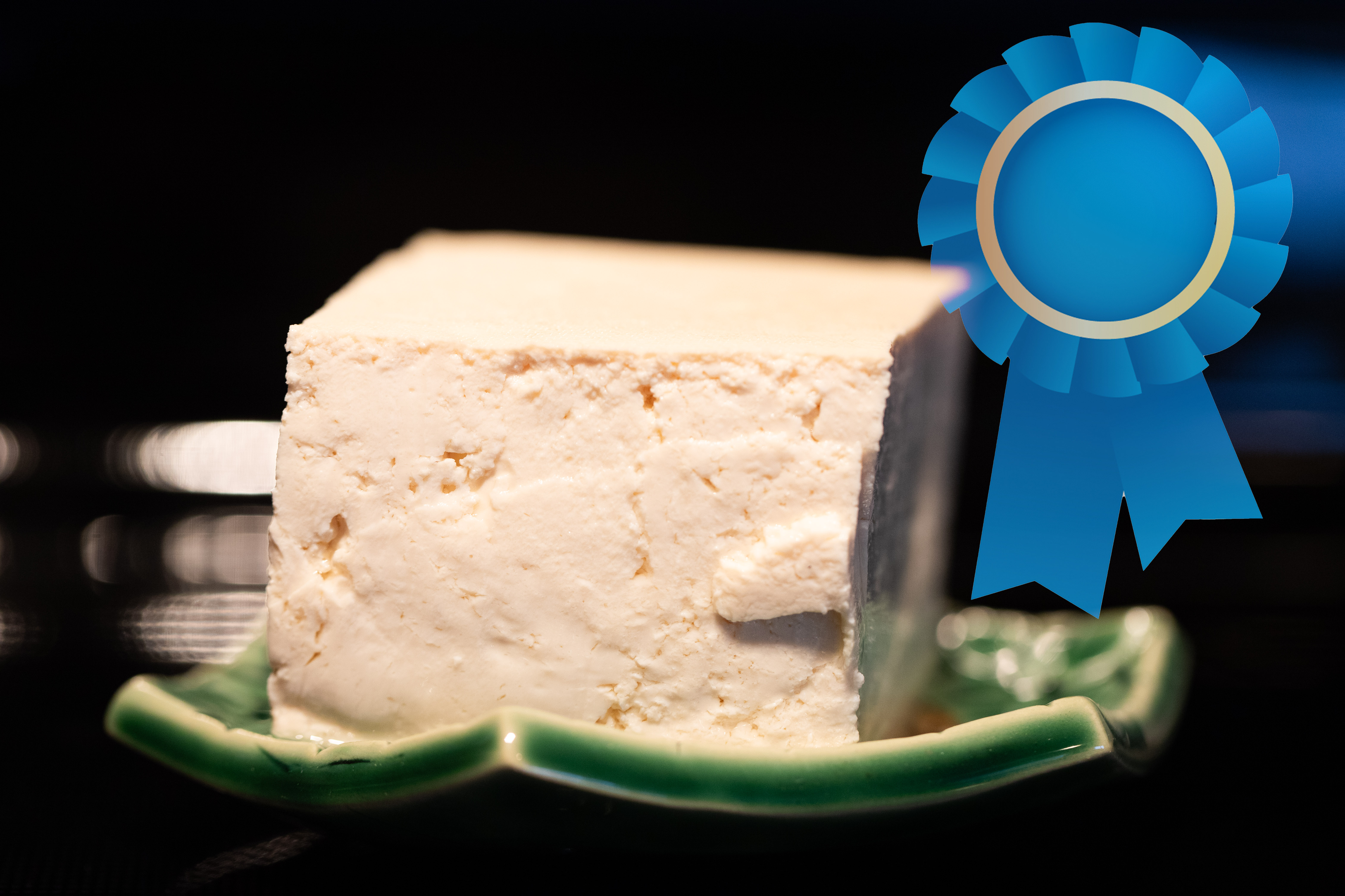 Close up of a block of tofu on a decorative plate with a blue ribbon icon overlaid to the right. Photo and processing by Tracy Isaacs. Blue Ribbon icon by Ikaika on Freepik.
