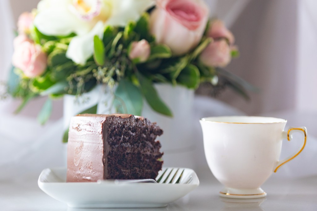 Chocolate layer cake on a square plate, china cup beside, and a flower arrangement blurred a bit in the background. Photo by Tracy Isaacs.
