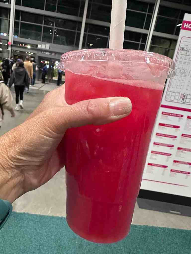 Tracy's hand holding a plastic take-out drink cup filled with watermelon drink, thick paper straw, people in the background. Photo by Tracy Isaacs