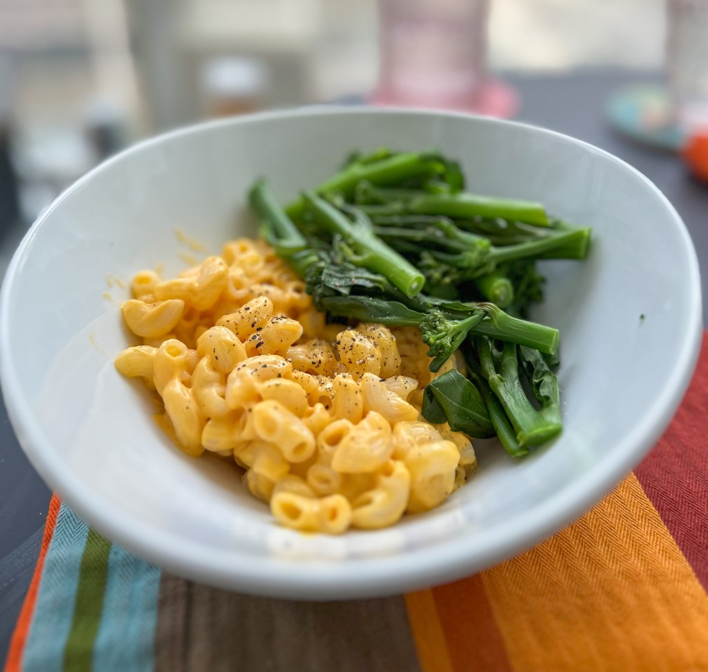 Round white bowl with half Daiya dairy-free cheddar mac and cheese and half young broccoli, steamed, sitting on a linen place mat, blurred window scene in the background. Photo by Tracy Isaacs