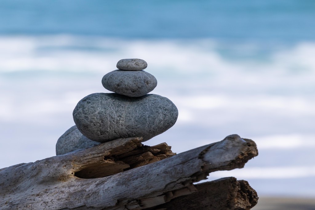 Four rocks stacked on a piece of driftwood on a beach, sea and surf in the background. Photo by Tracy Isaacs