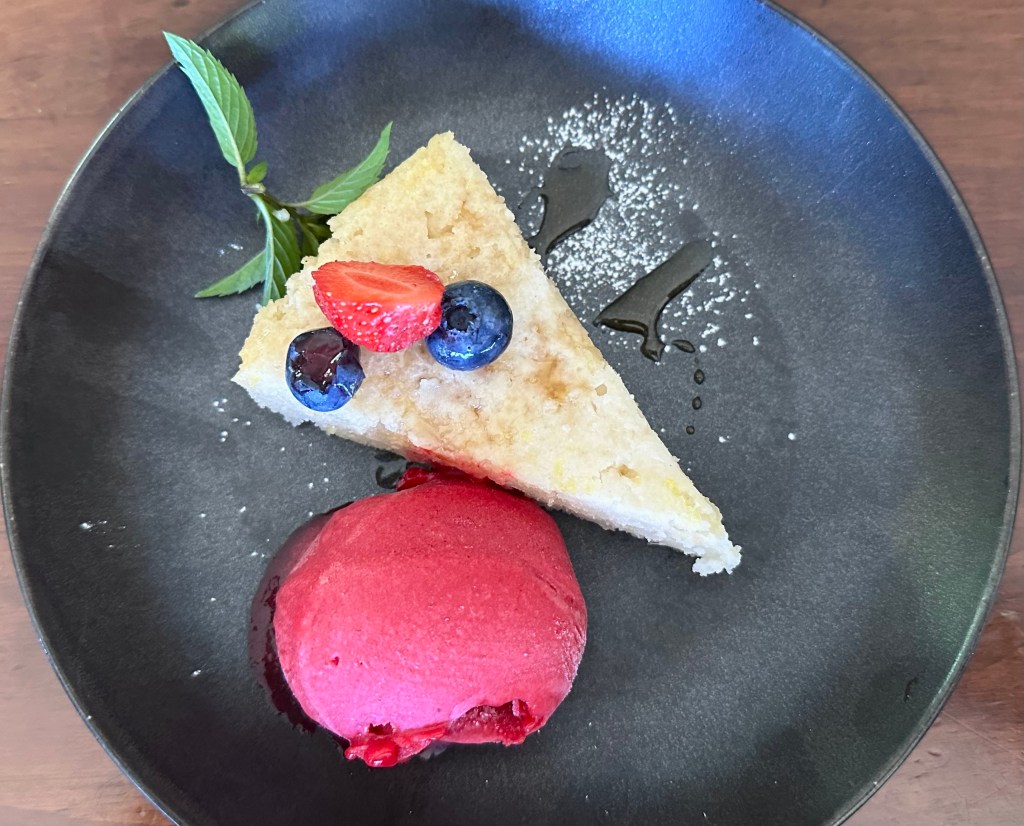 Vegan lemon cake topped with berries and fresh mint, scoop of raspberry sorbet beside, served on a dark round plate.