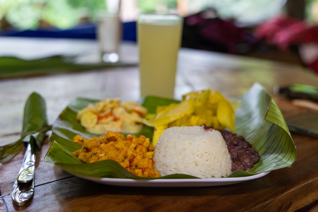Mixed plate of banana salad, plantain chips, salsa, rice, and beans on a banana leaf, glass of soursop juice in background. Photo by Tracy Isaacs