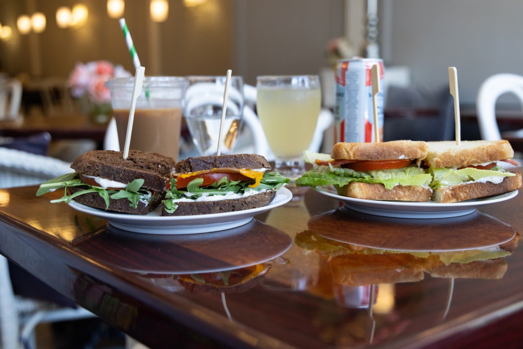 Interior of the Vegan Danish Bakery with a close-up of two plates with sandwiches on them, on a shiny wood table, drinks and lights in the background. Photo by Tracy Isaacs