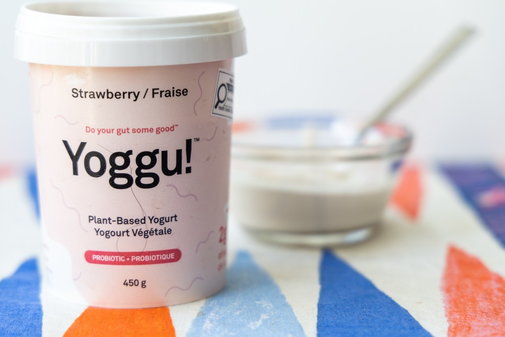 Container of strawberry Yoggu! in the foreground and a small glass bowl of yogurt with a spoon in it in the background. It's all on a tea-towel with a triangular print that looks like part of a backgammon board. Photo by Tracy Isaacs