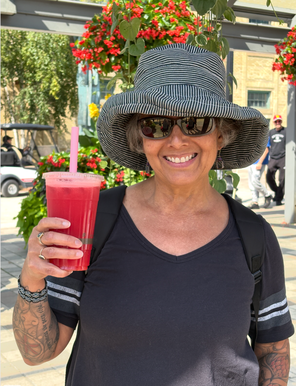 Image description: smiling woman (Tracy) in a wide brimmed hat, sunglasses, and a black t-shirt, holding up a watermelon drink. Photo by Diane.