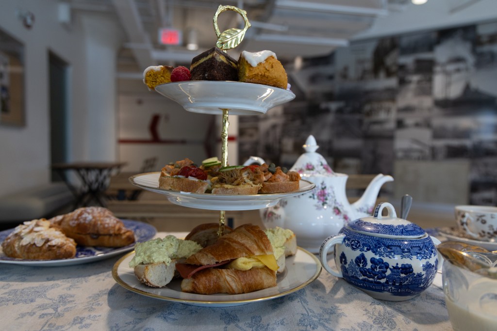 Image description: a three-tiered serving plate of Animal Liberation Kitchen's vegan tea. Bottom tier of vegan breakfast croissant sandwich and open faced tofu "egg" salad, middle tier of assorted open faced sandwiches, top tier of scones, nanaimo bars, berries. In background a china teapot, china lidded sugar bowl, china tea cup. Photo by Tracy Isaacs